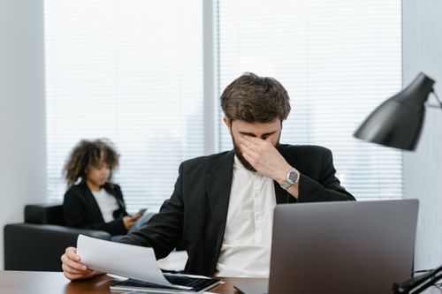 Free Businessman in an office covering his eyes while working on a laptop, feeling stressed. Stock Photo