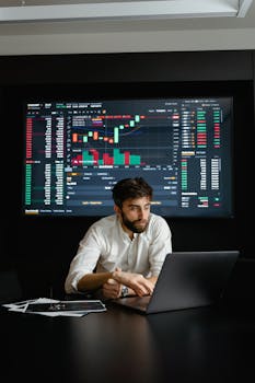 A man analyzing stock market data on a laptop in an office setting with a projector screen displaying financial charts.