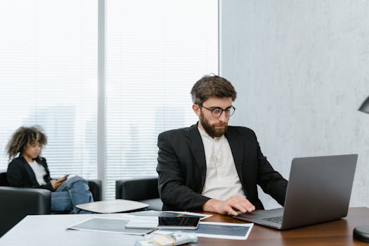 Man in black suit focusing on laptop, woman in background. Modern office setting.