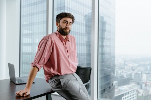 Free A businessman with facial hair in a pink shirt sits by a large window in a modern office building. Stock Photo