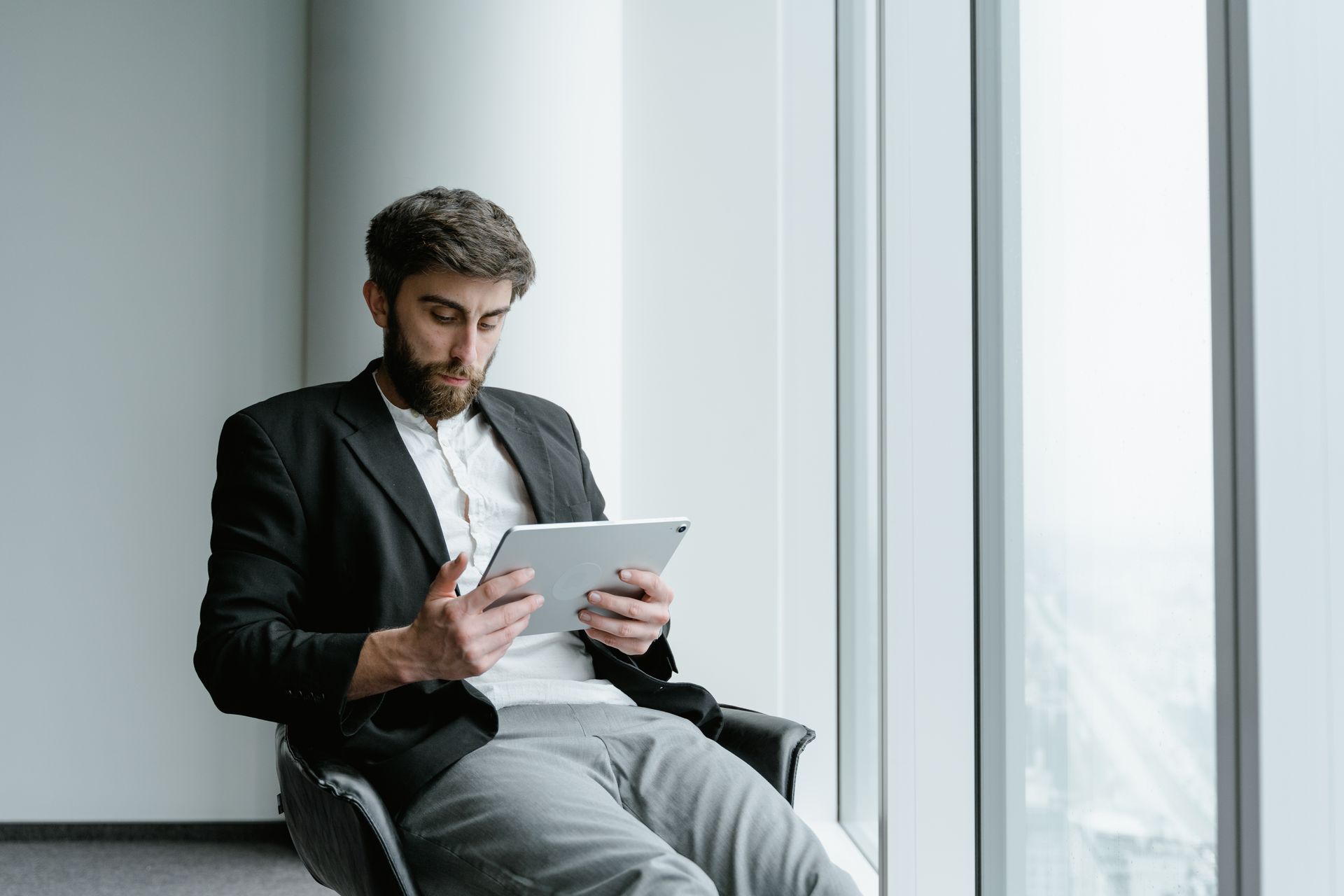 A man in formal attire working on a tablet while seated by a large office window.