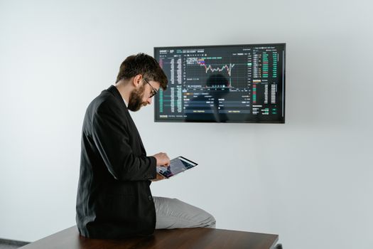 Young man in office analyzing financial graphs on a tablet with stock market screen displayed.