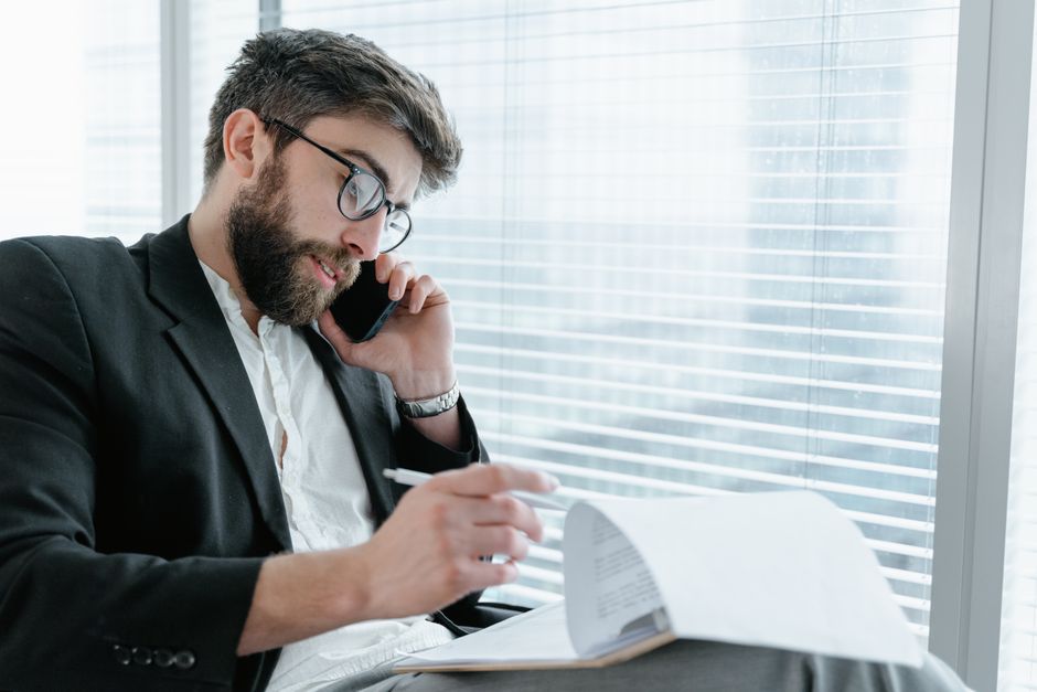 Hombre de negocios con gafas haciendo una llamada telefónica mientras revisa documentos en un entorno de oficina moderno.