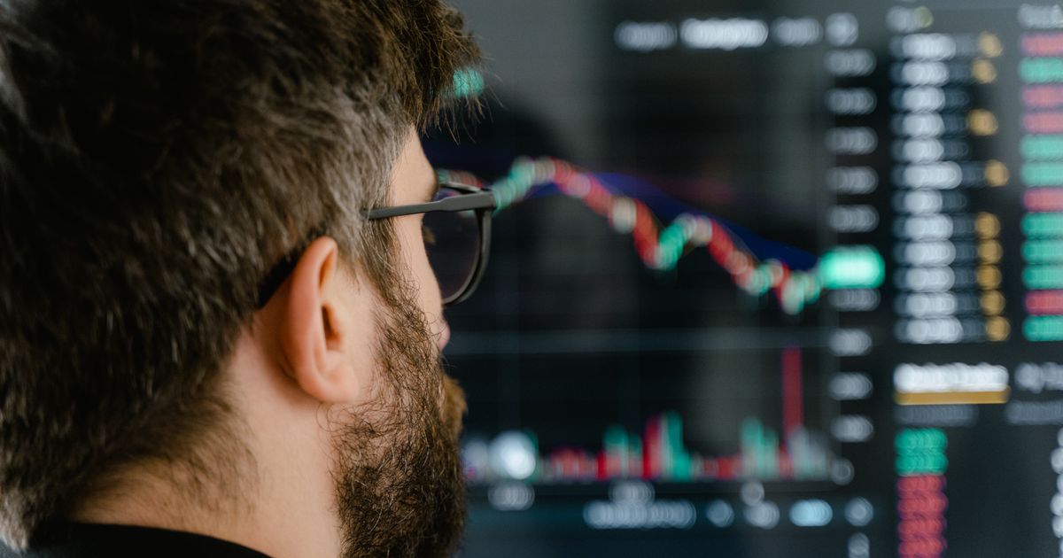 A bearded man with glasses examines trading charts on a computer screen, indicating financial analysis.