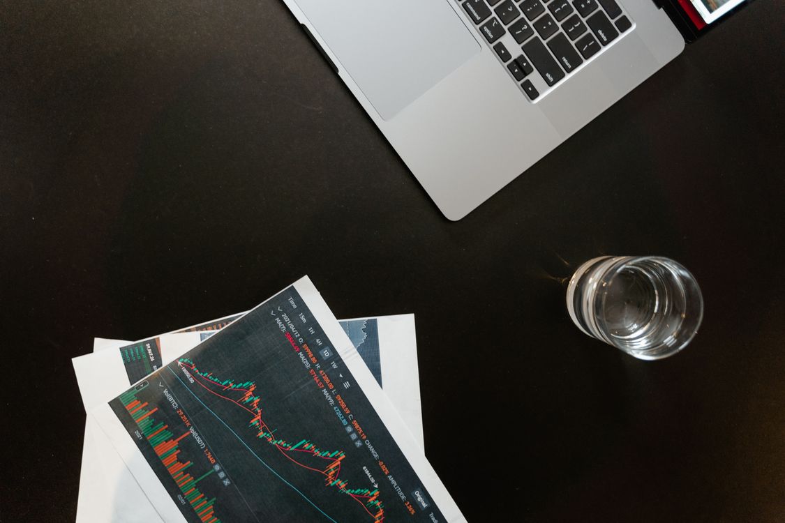Free Professional workspace featuring financial graphs, laptop, and glass of water on a sleek desk. Stock Photo