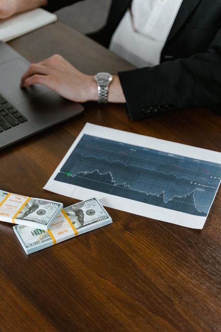 Photo by Tima Miroshnichenko A desk scene with cash bundles, a chart printout, and a person working on a laptop.