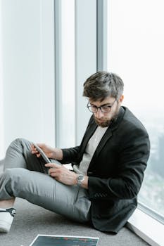 A bearded man in a black blazer sits by a window analyzing data on a tablet.