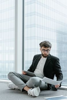 Young businessman in formal attire reading papers by a large glass window, emphasizing concentration.