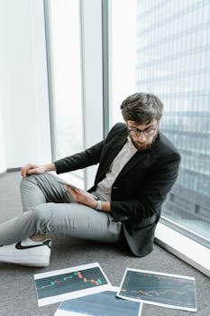 Man in black blazer and eyeglasses analyzing financial papers sitting by a glass window.