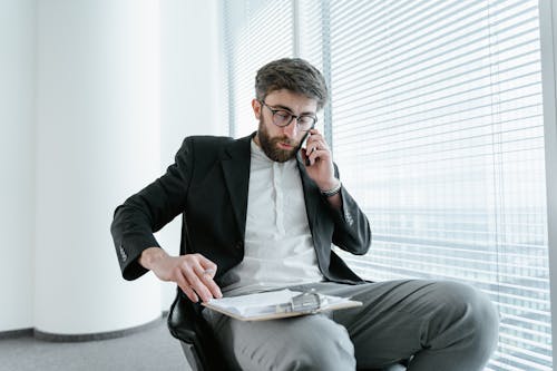 Free A Man in Black Suit Sitting on the Chair while Talking on the Phone Stock Photo