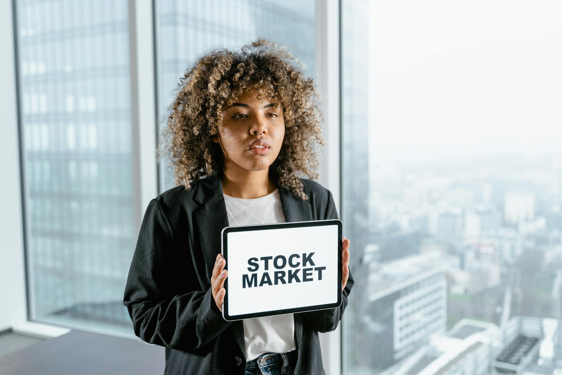 Woman in office holding a tablet with 'Stock Market' displaying on the screen, cityscape background.