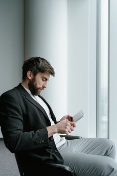 Bearded man in black blazer sits by window, using tablet in corporate setting.