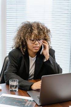 Businesswoman with curly hair talking on phone while working on laptop indoors.