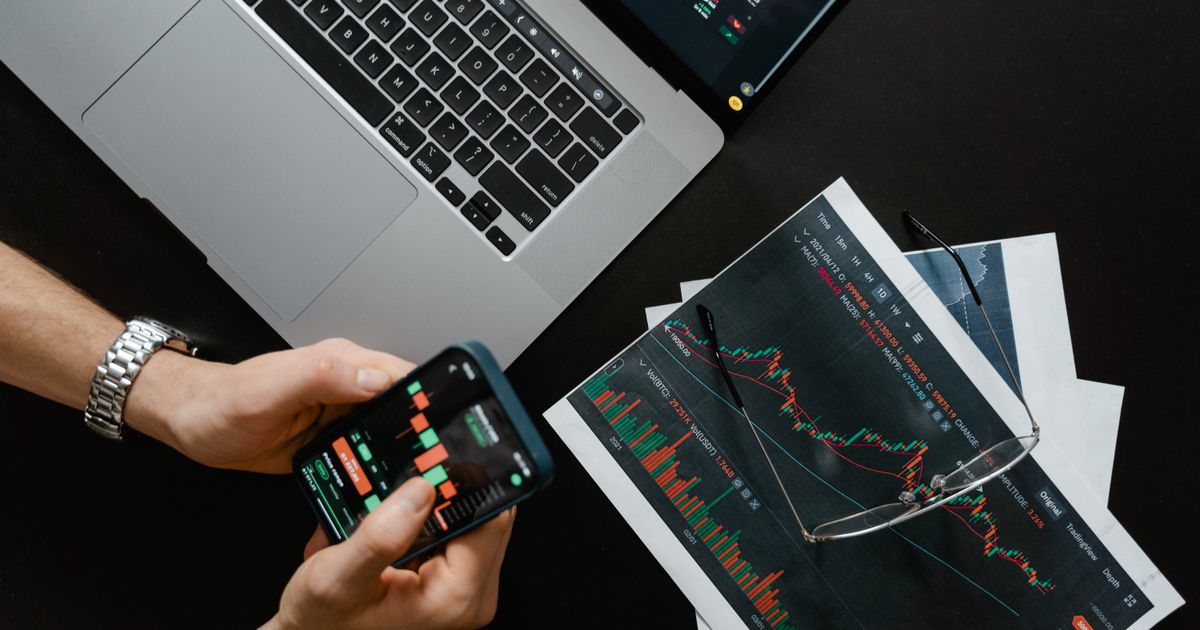 Manos sosteniendo un celular con datos financieros junto a una laptop con gráficas del mercado para comprar acciones en México desde una app