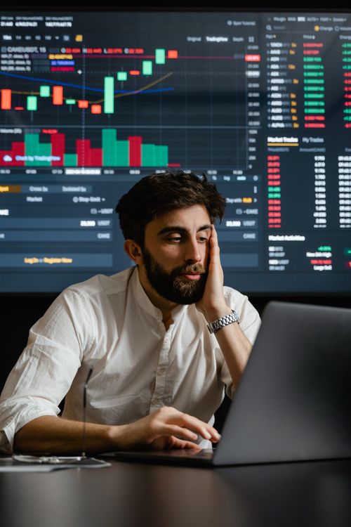 Free Bearded businessman analyzing financial statistics on a laptop, with stock market graphs in the background. Stock Photo