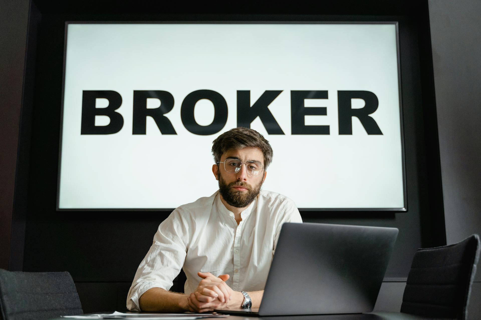 A broker with glasses sits confidently in front of a laptop in a modern conference room.