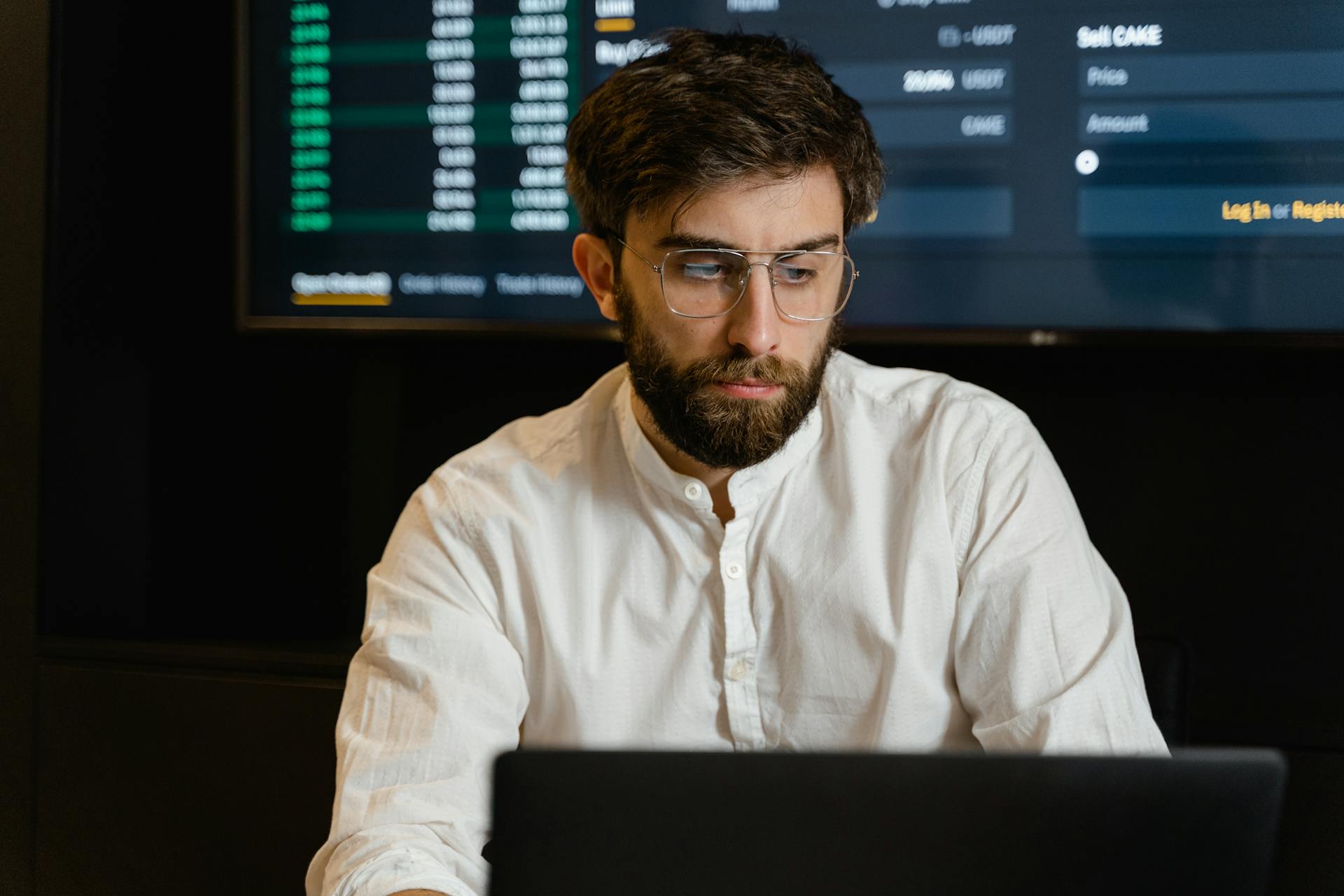 Man with glasses and beard focused on his laptop in a modern office setting with financial data on screen.