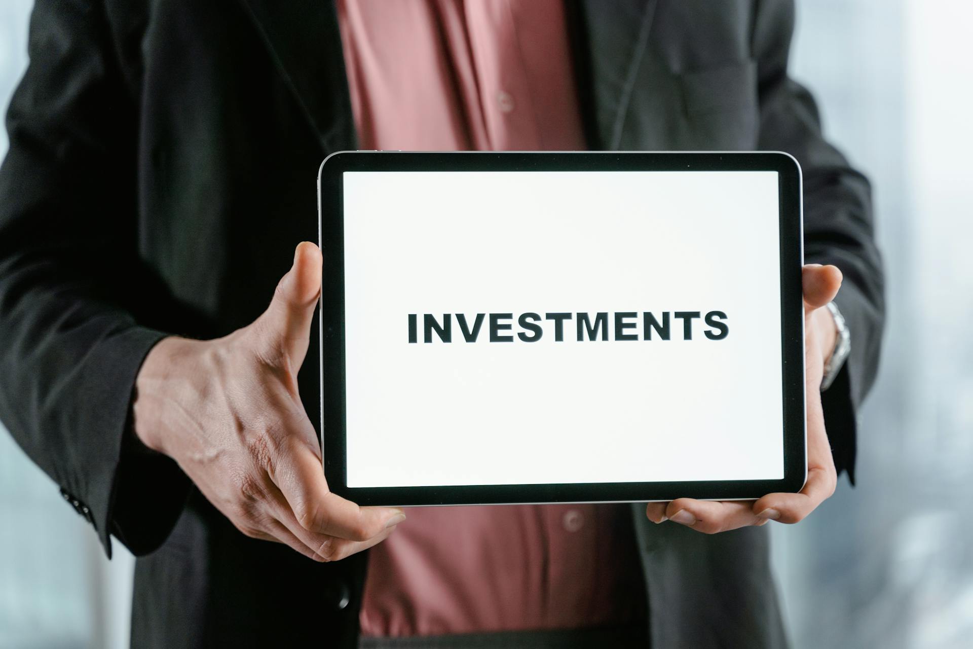 A businessman in a suit holds a tablet displaying the text 'INVESTMENTS'.