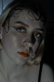 Confident young woman with bright creative makeup and hairstyle looking at camera in dark room
