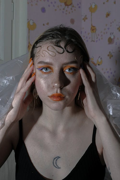 Calm young woman with creative bright makeup and hairstyle touching head and looking at camera in room