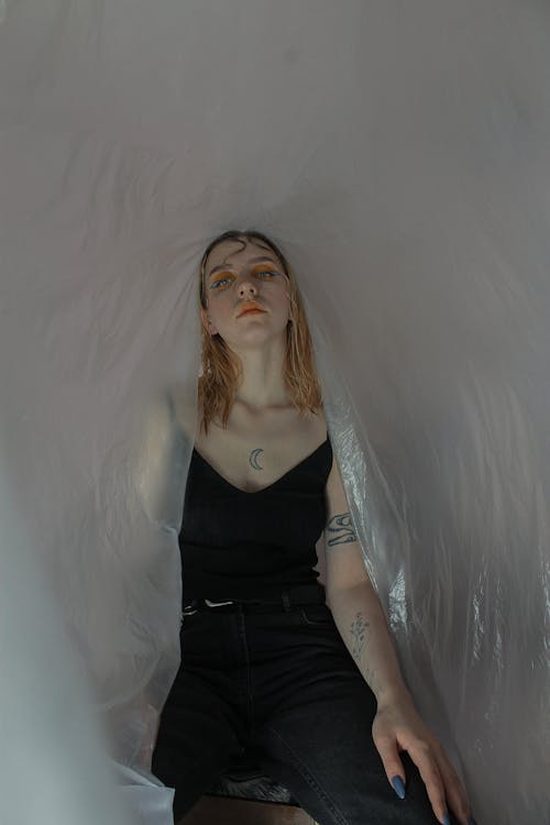 Free Melancholy female model in black outfit sitting among plastic wrap and looking away Stock Photo
