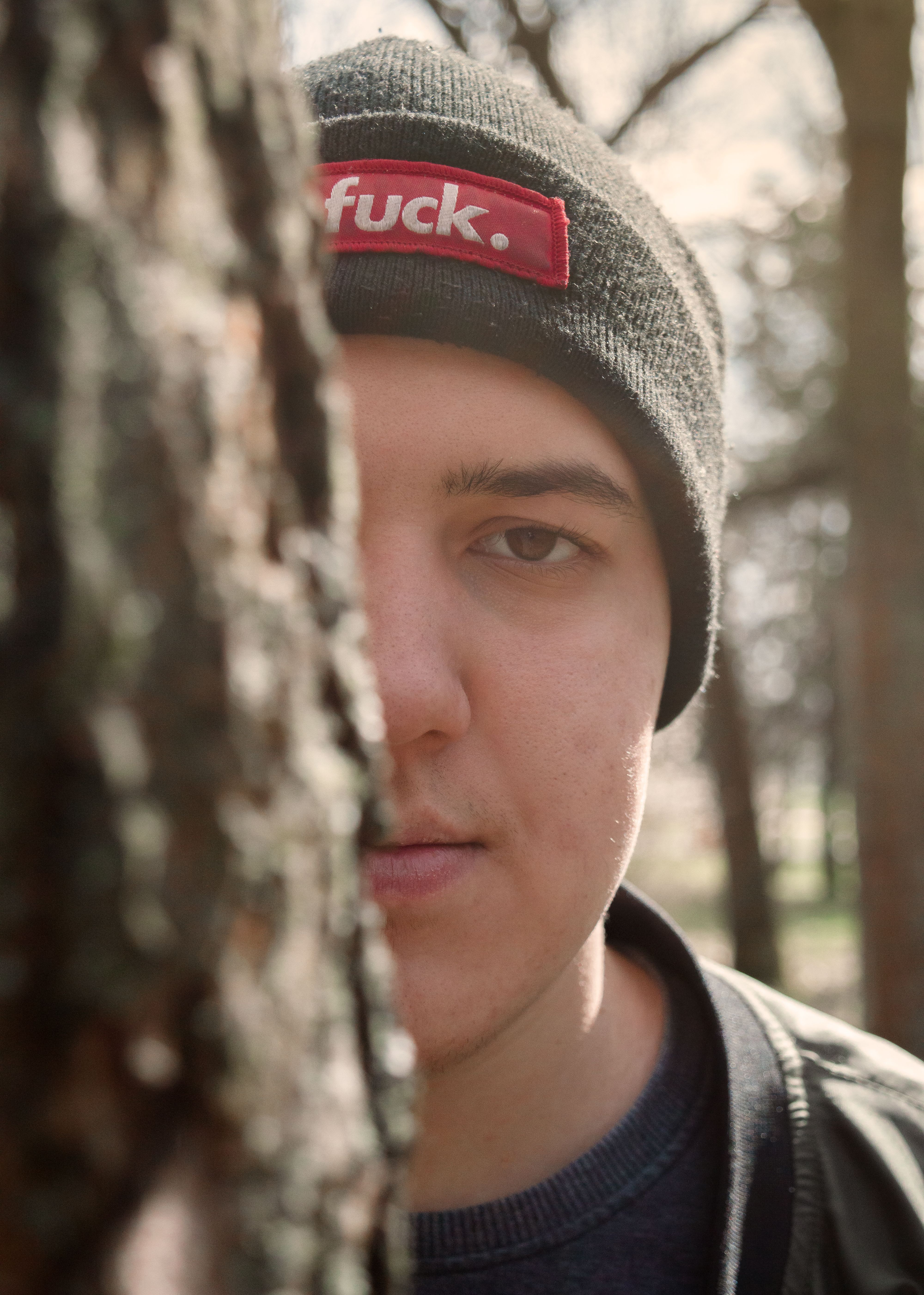 A young man wearing a beanie, partially obscured behind a tree in a park setting.