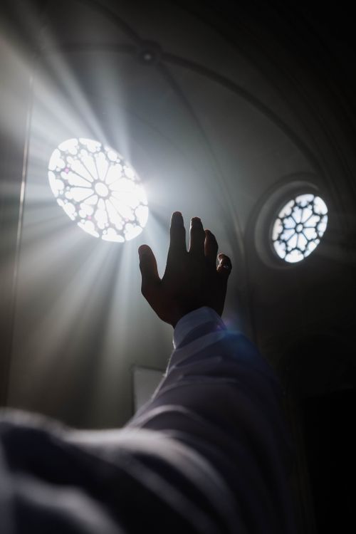 Free A hand reaches towards sunlight streaming through stained glass windows in a dimly lit room, creating a silhouette. Stock Photo