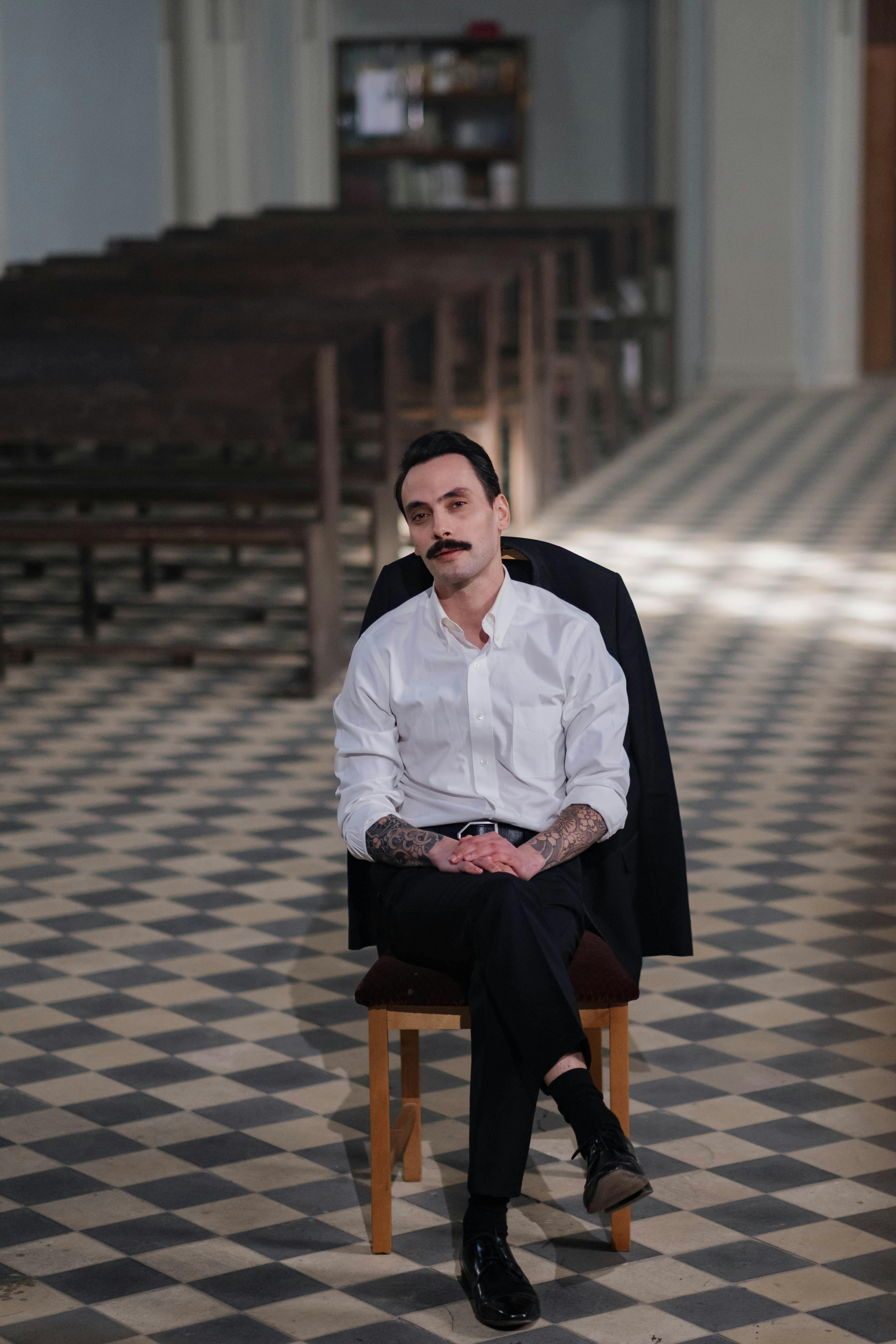 Man with tattoos and mustache seated on wooden chair in church with checkered floor.
