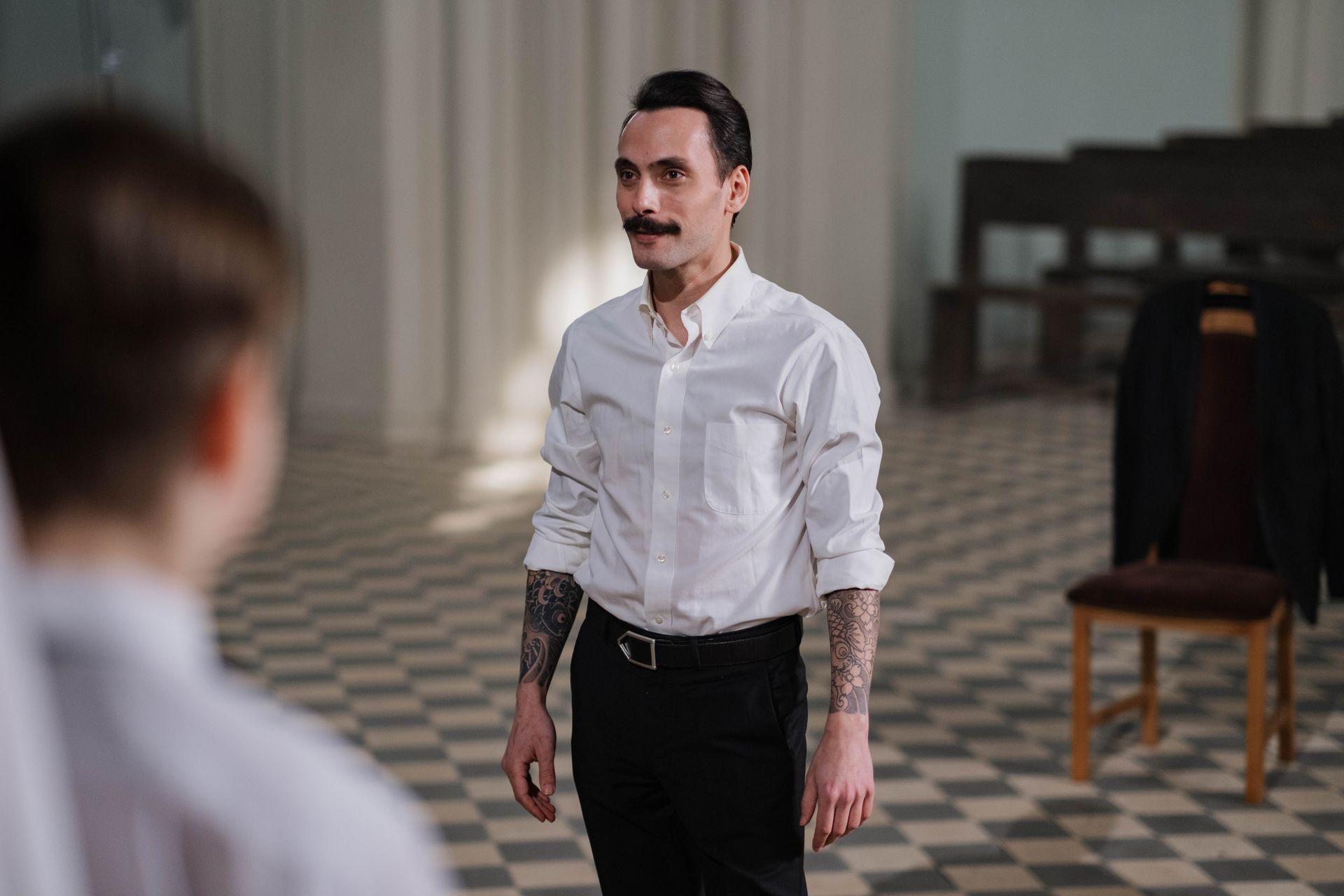 A man with tattoos and a moustache leads a practice indoors, wearing a white shirt.