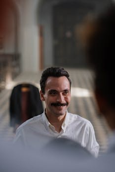 Portrait of a smiling man with a moustache and white shirt inside an elegant building.