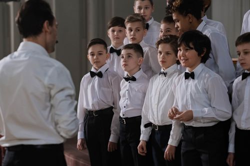 Free Boys choir dressed elegantly in white shirts and bow ties, led by a conductor indoors. Stock Photo