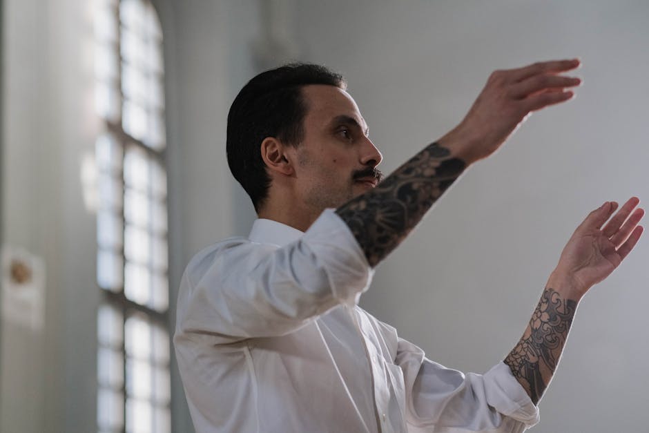 A tattooed conductor in white guiding musicians indoors during a musical performance.