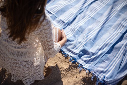 Free A woman in lace sits on a sandy Portuguese beach, beside a blue striped blanket, enjoying a sunny day. Stock Photo
