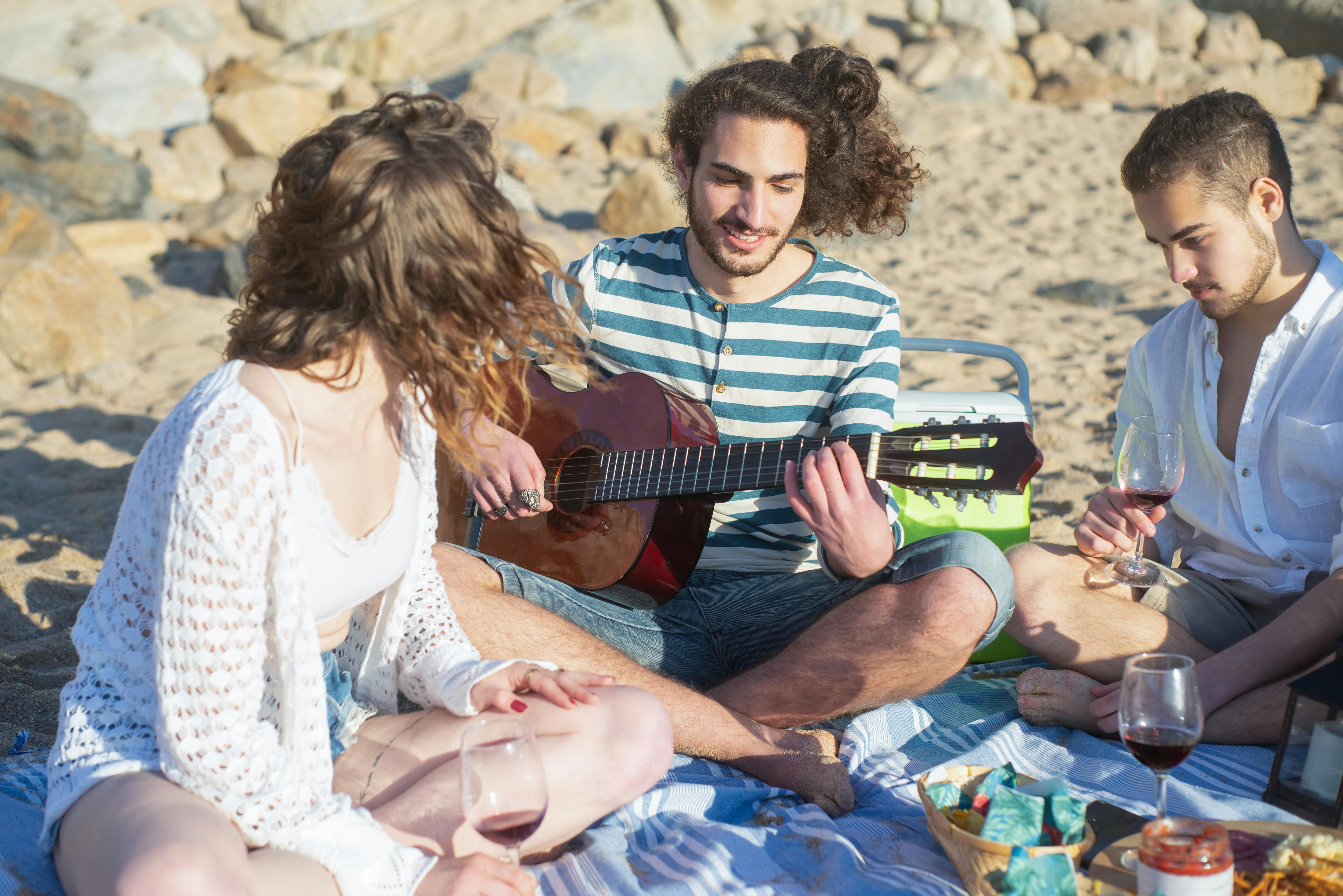 a man playing guitar with his friends while sitting on a picnic blanket