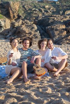 A cheerful group of young adults having a beach picnic in Portugal. Smiling and relaxing with a ukulele and drinks on a sunny day.