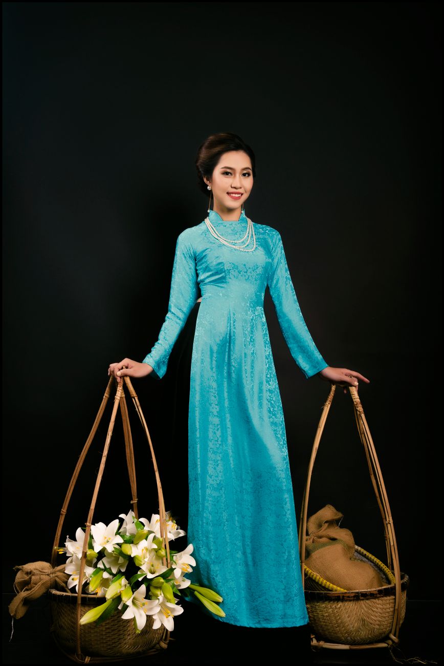 Portrait of an Asian woman in blue ao dai holding woven baskets against a black background.