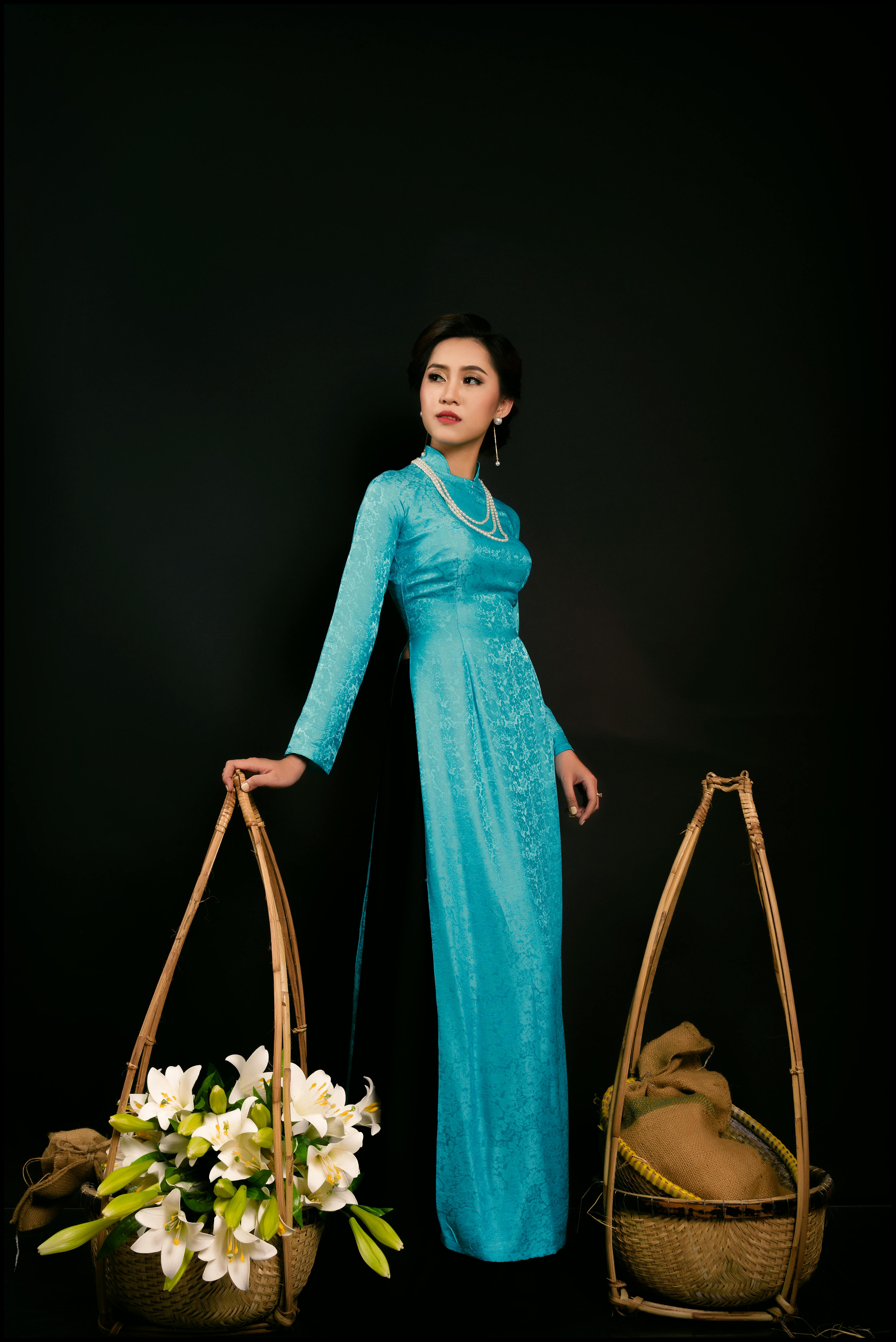 Asian woman in a traditional blue ao dai holding woven baskets with flowers against a black background.