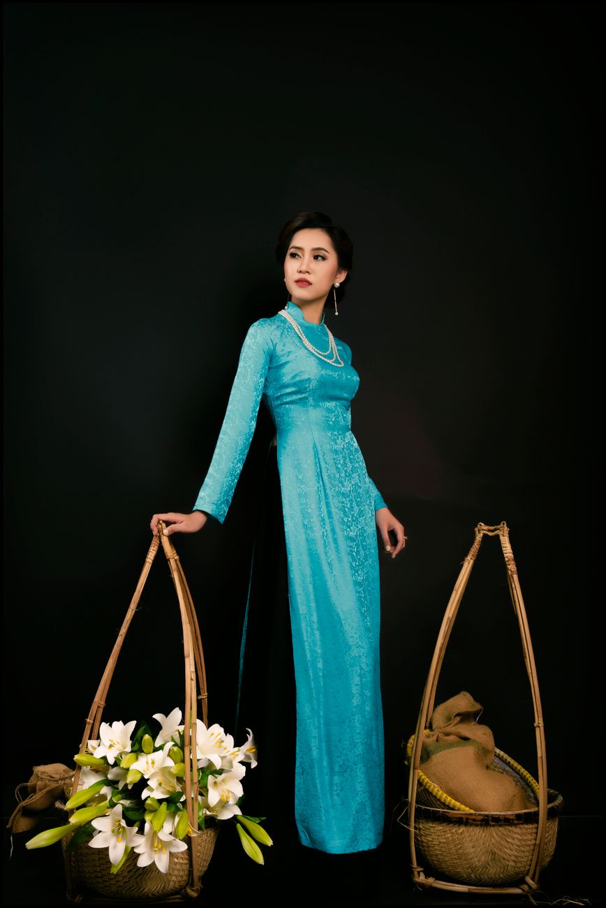 Asian woman in a traditional blue ao dai holding woven baskets with flowers against a black background.