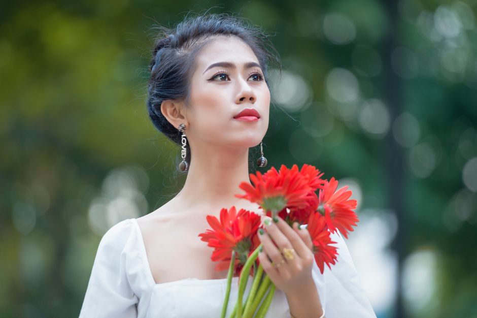 A woman in a white dress holds red flowers, gazing into the distance outdoors.