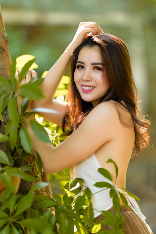 Young sensual ethnic female with long brown wavy hair and bare shoulders wearing white top and skirt leaning on tree and looking at camera in park