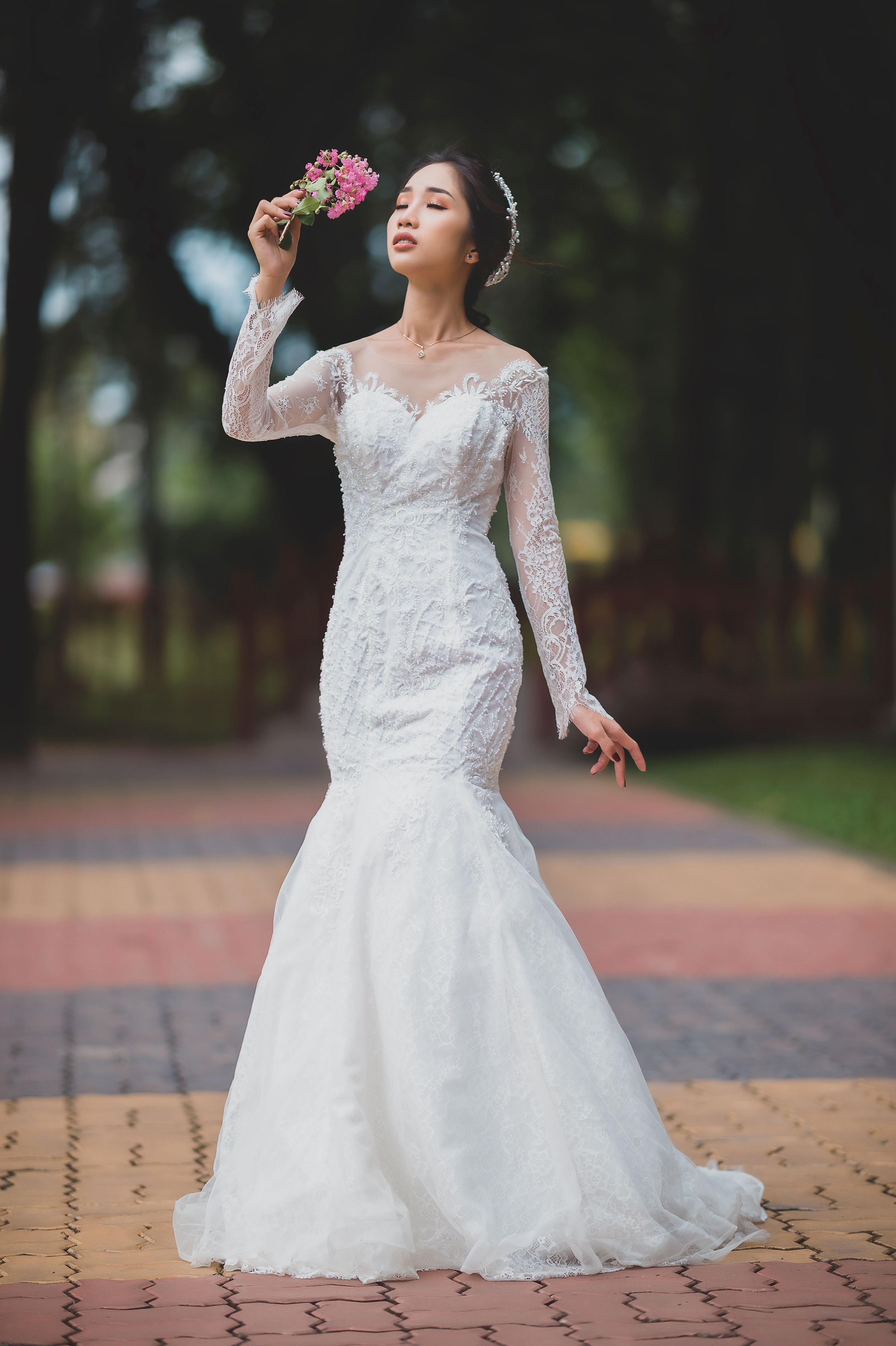 Full body of charming young Asian female with closed eyes and long dark hair wearing white wedding dress standing on walkway in green park with blooming flowers