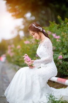 Beautiful bride in lace wedding dress, sitting outdoors with pink flowers, exuding elegance and serenity.
