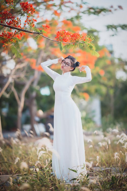 A Woman in White Dress while Wearing an Eyeglasses Standing Near the Tree with Blooming Flowers