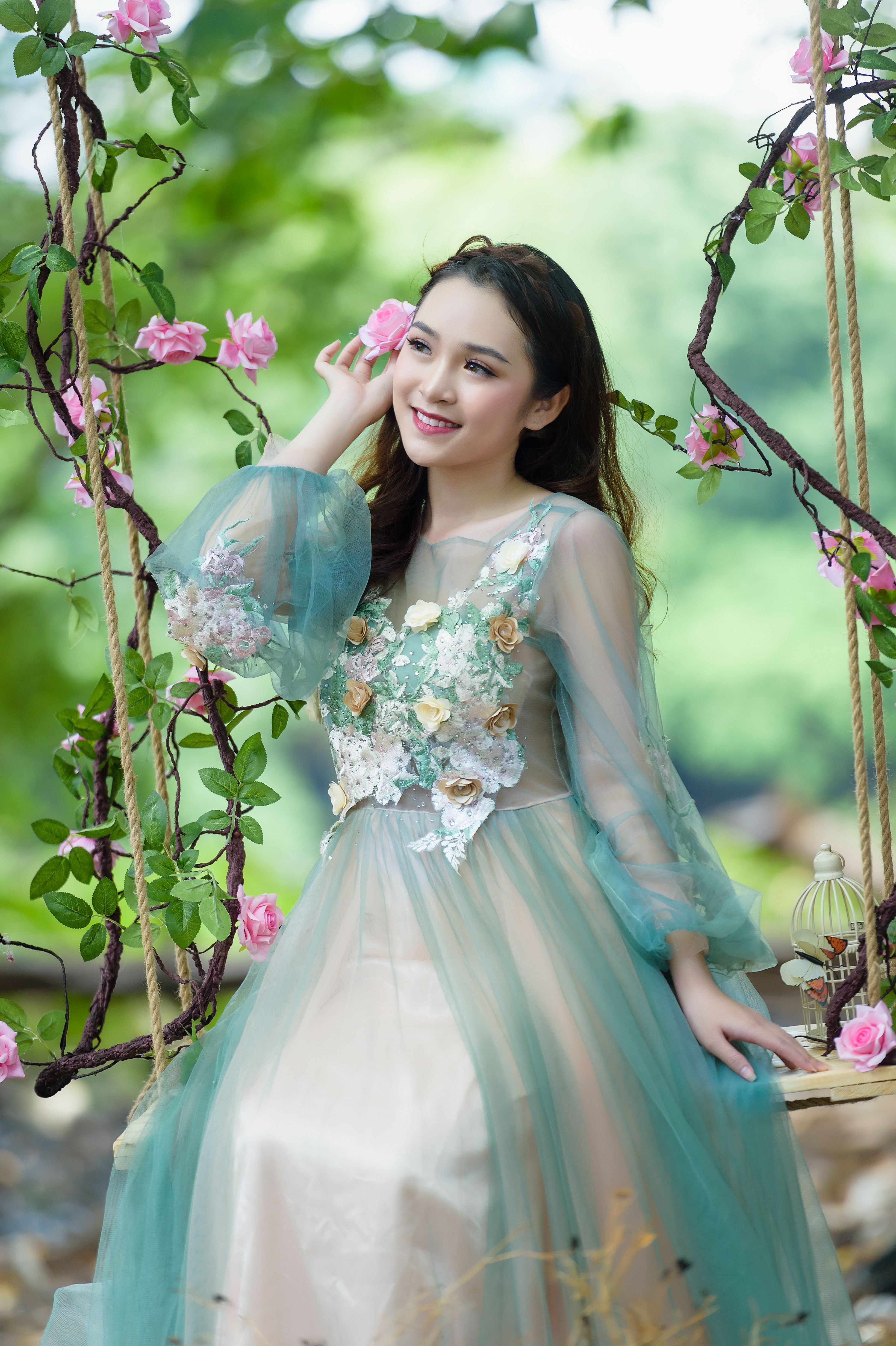 A smiling woman in an elegant floral dress sits on a decorated swing outdoors.