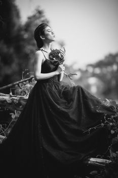 Graceful woman in black evening gown holding a rose, captured in a serene, monochrome outdoor setting.