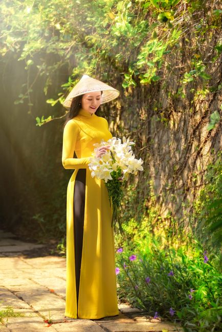 A woman in a traditional yellow Ao Dai, holding a white lily bouquet, stands outdoors, surrounded by lush greenery.