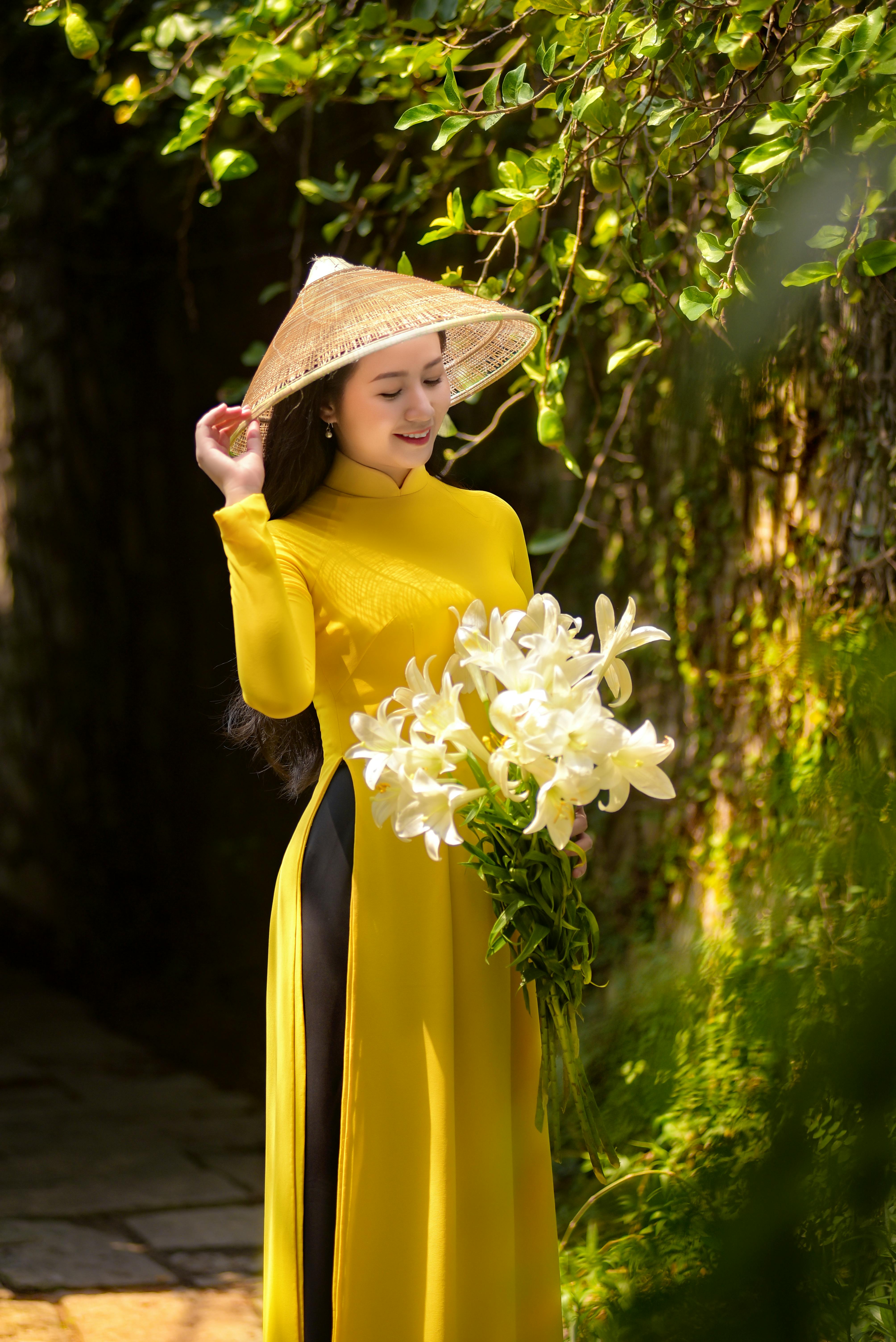 A woman in a yellow traditional dress and conical hat holding white lilies outdoors.