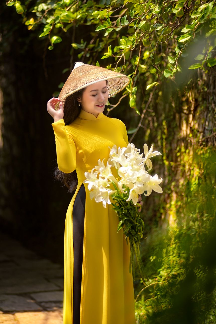 A woman in a yellow traditional dress and conical hat holding white lilies outdoors.