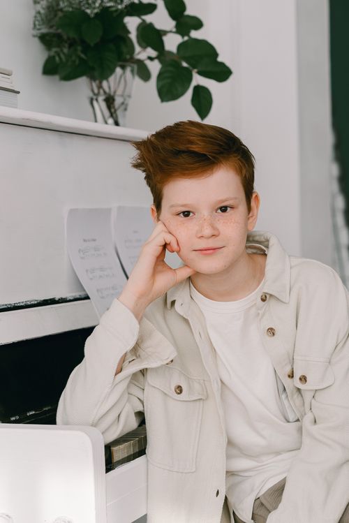 Free Red-haired boy leaning against a piano, looking at the camera with a soft smile. Stock Photo