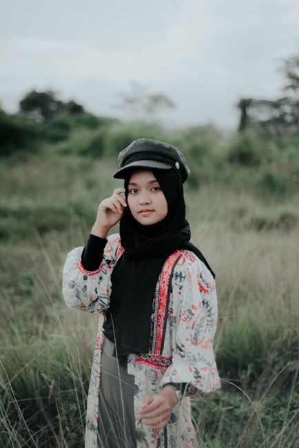 A young woman in a hijab stands confidently in a grassy field wearing a unique outfit and cap.